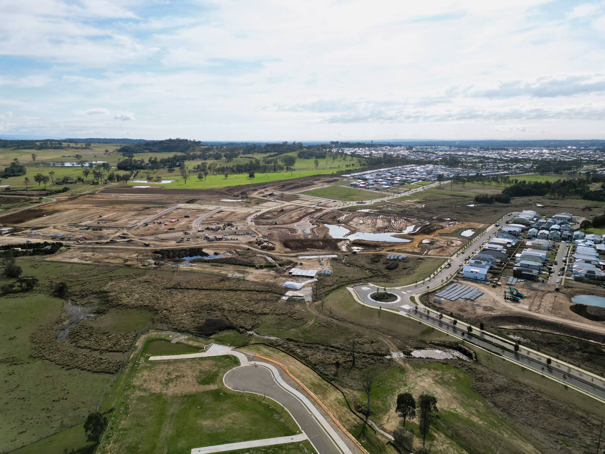 Aerial view of a large construction site with cleared land, dirt roads, and scattered equipment, adjacent to a residential area and green fields under a partly cloudy sky.