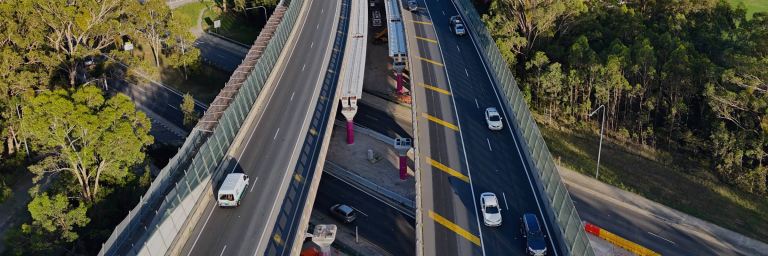 Aerial view of a multi-lane highway interchange surrounded by dense green vegetation. Some lanes are under construction with visible construction vehicles and barriers, while others have moving traffic. Urban development is visible in the distance beyond the forested area.