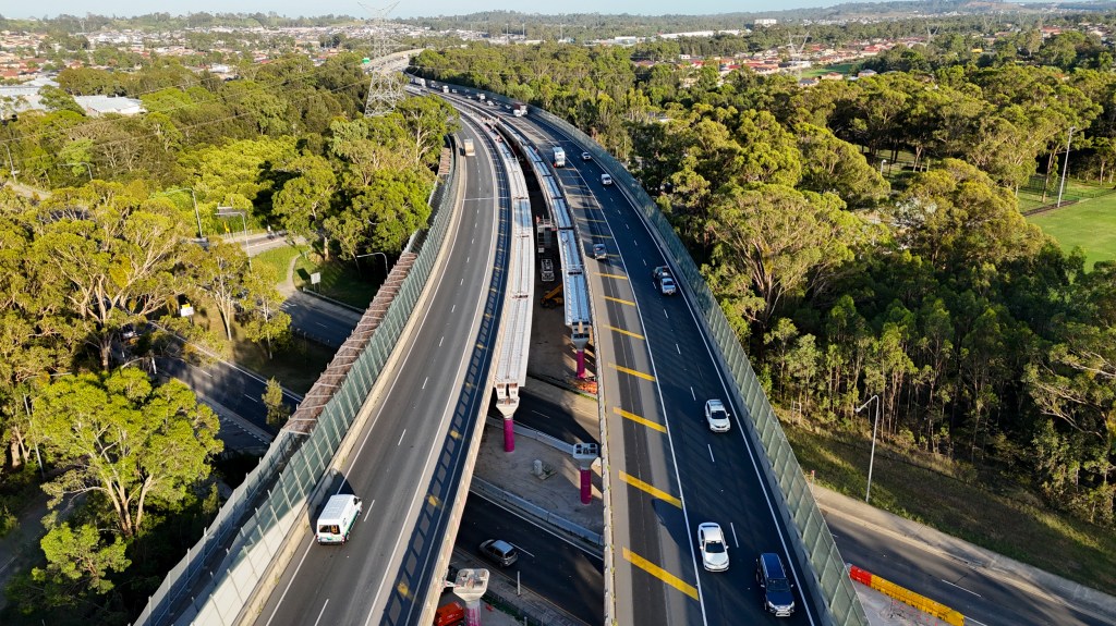Aerial view of a multi-lane highway interchange surrounded by dense green vegetation. Some lanes are under construction with visible construction vehicles and barriers, while others have moving traffic. Urban development is visible in the distance beyond the forested area.