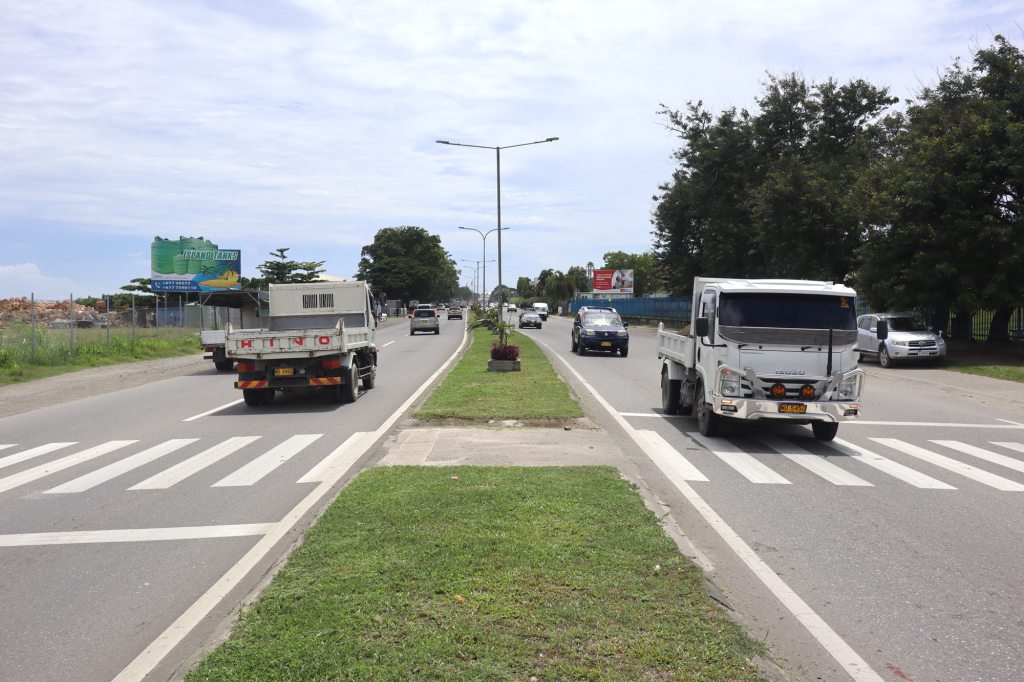 Dual carriageway road in the Solomon Islands with vehicles travelling in both directions, featuring pedestrian crossings and central median planting