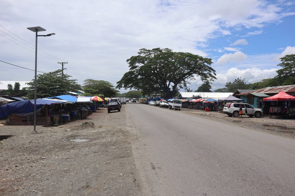 Roadside market area along a paved road in the Solomon Islands with parked vehicles and small stalls on both sides