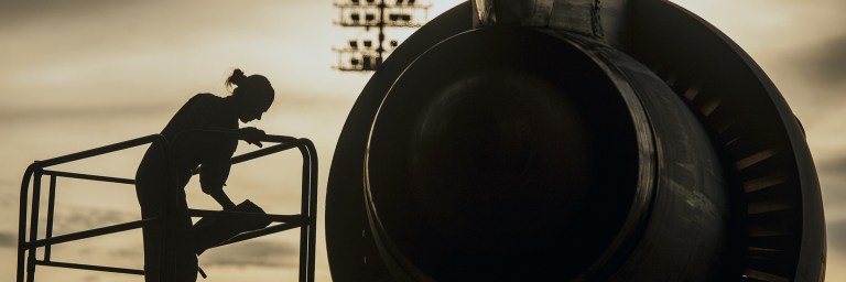 Aircraft mechanic kneels beside an aircraft engine or intake, appearing to inspect or adjust components.