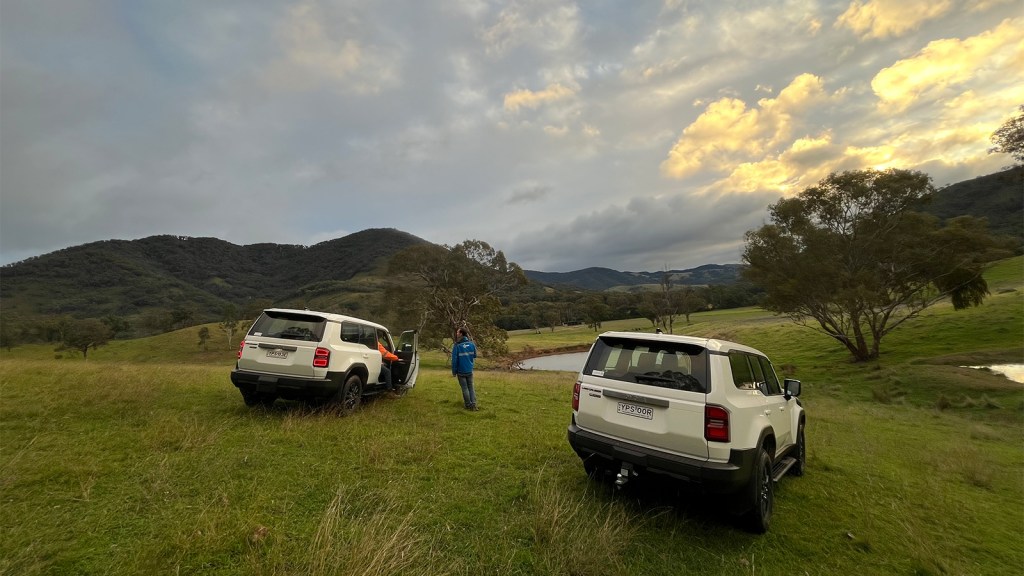 Two SUVs parked on grassy hills with a person beside an open door under a cloudy sunset sky.