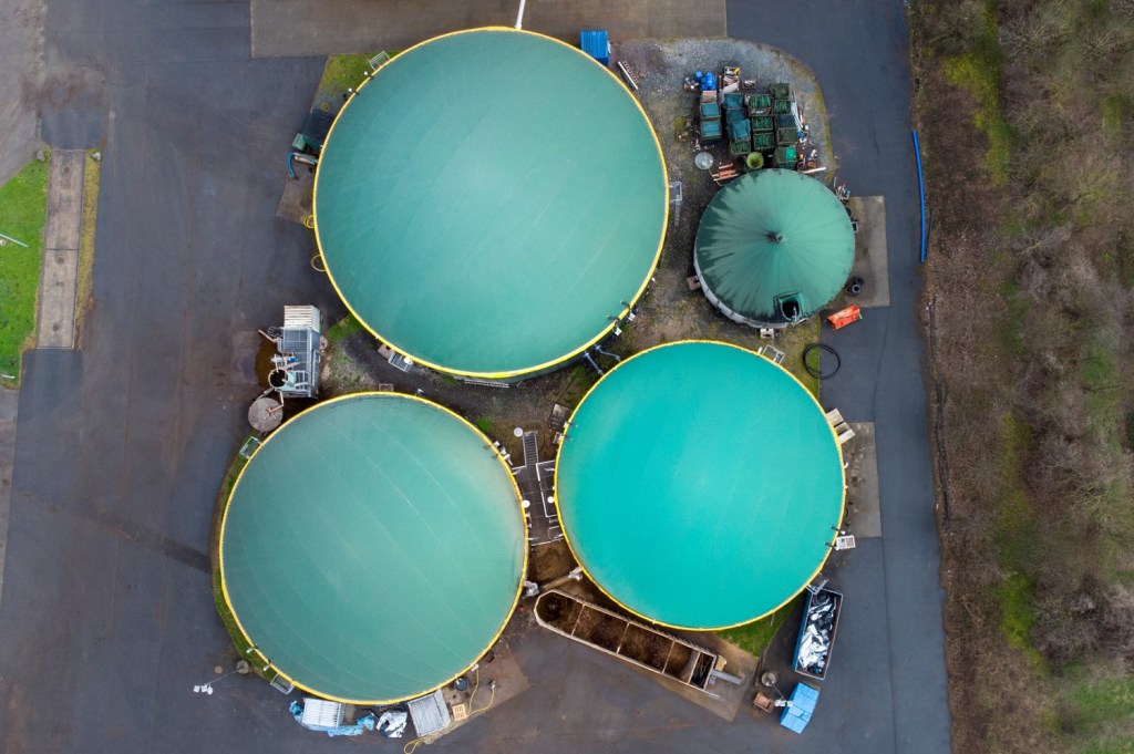 Aerial view of a biogas plant featuring three large green digesters, a smaller storage tank, and surrounding industrial infrastructure.