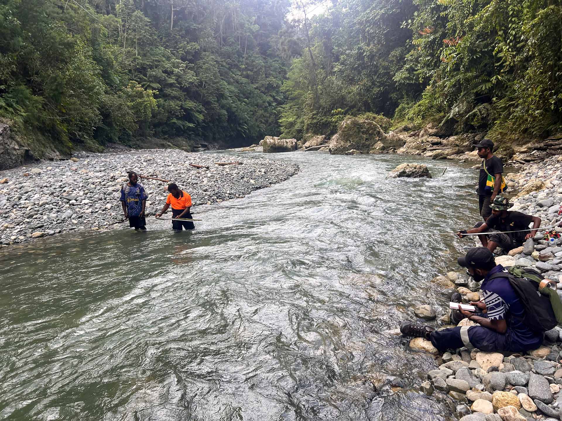 Field team measuring a river in a forested area, with several people collecting data along the rocky riverbank and in the water.