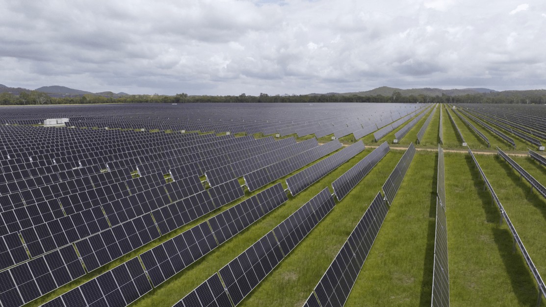 Aerial view of a large solar farm with rows of dark solar panels stretching across a green landscape.