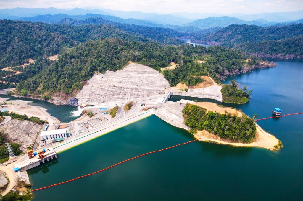 Aerial view of a hydroelectric dam and reservoir surrounded by forested mountains and clear blue water.