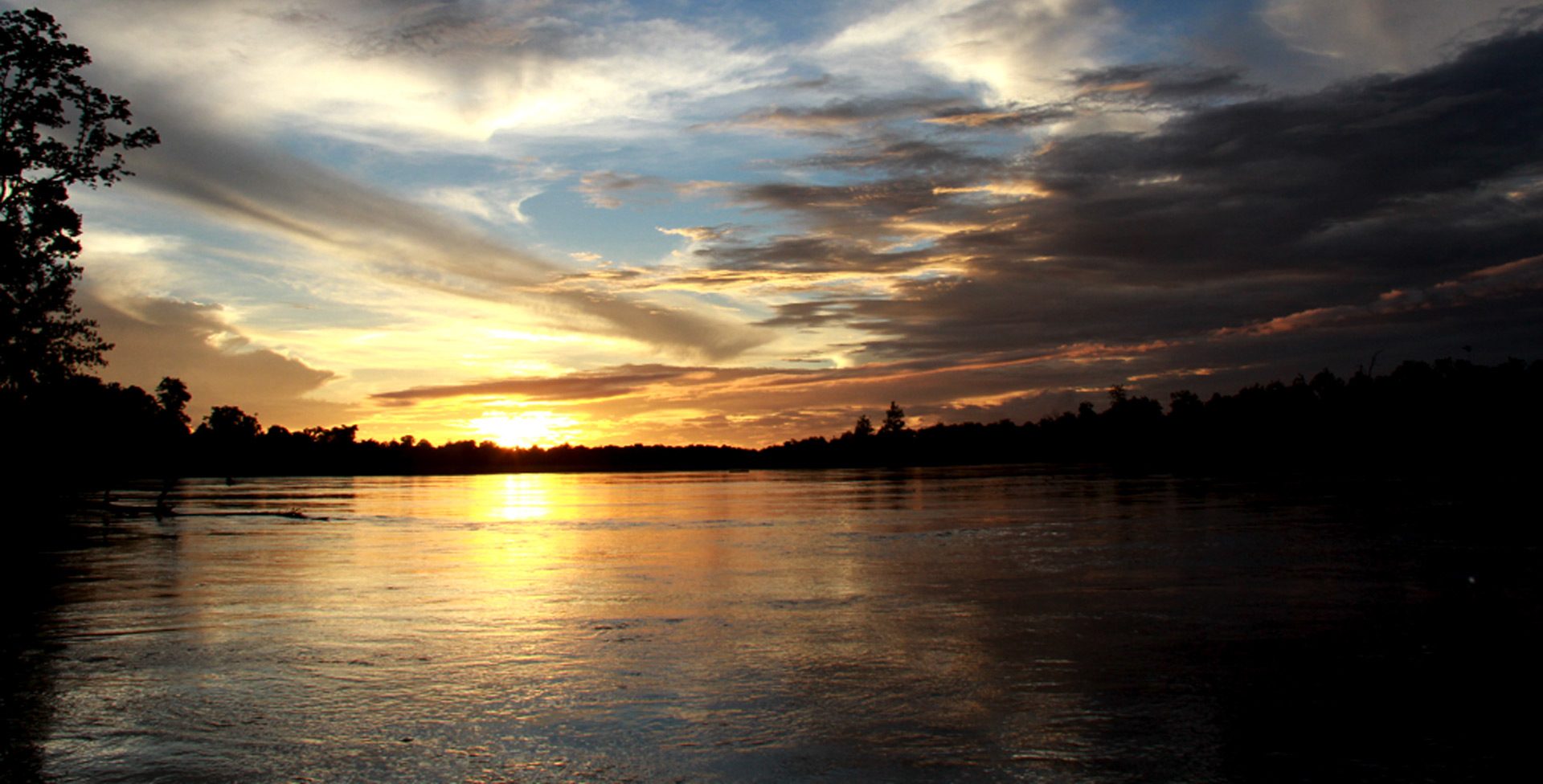 Golden sunset over a river with dramatic cloud formations and tree silhouettes along the horizon.