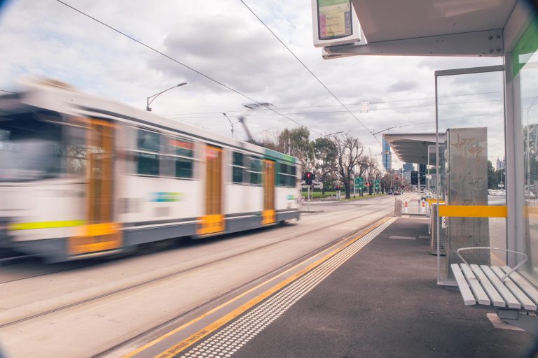 A tram passes through Hoddle Street at a modern tram stop, with motion blur showing movement along the tracks.