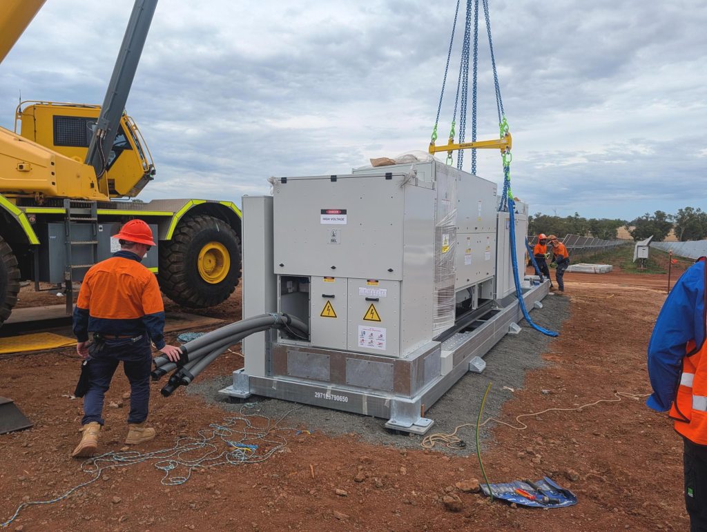 Workers install battery energy storage equipment at the Maryvale Solar and Energy Storage Project, using a crane to position a large electrical unit alongside solar infrastructure at the site.