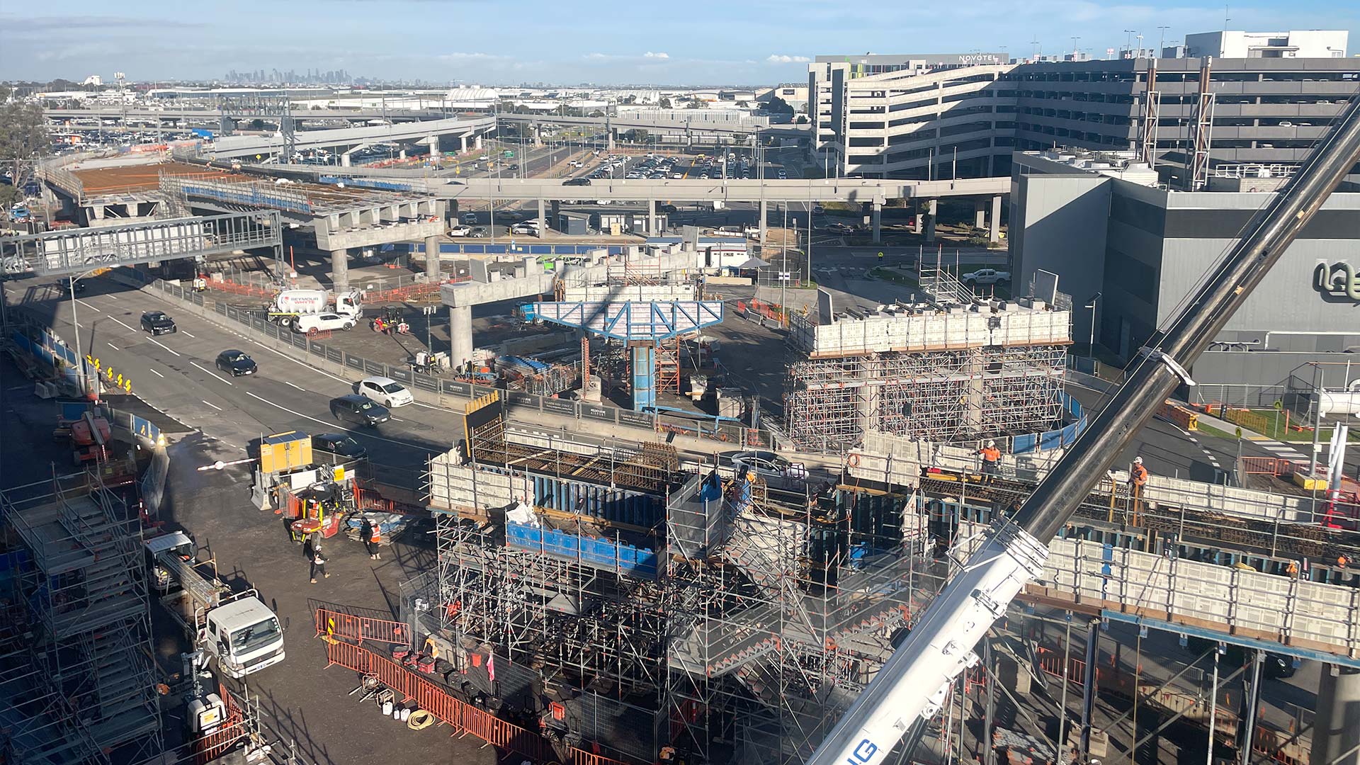A wide view of Melbourne Airport showing major construction works near the terminal. Multiple elevated roadways, concrete bridge structures, scaffolding, and a large crane dominate the foreground, while vehicles travel along access roads below. Airport buildings, parking facilities, and terminal infrastructure are visible in the background under a clear daytime sky.