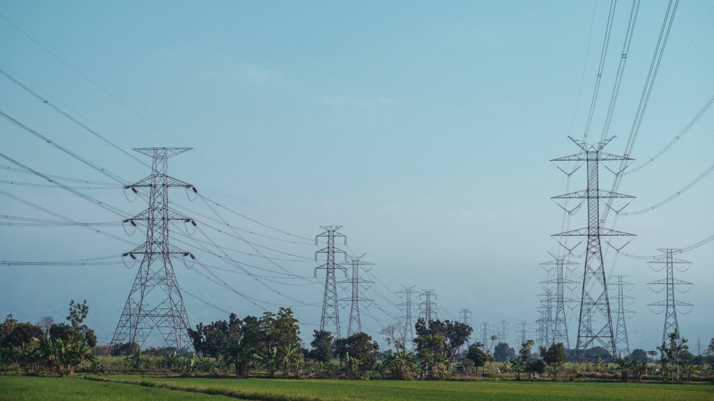 Copilot said: Electrical transmission towers and power lines stretching across a green agricultural landscape under a hazy blue sky.