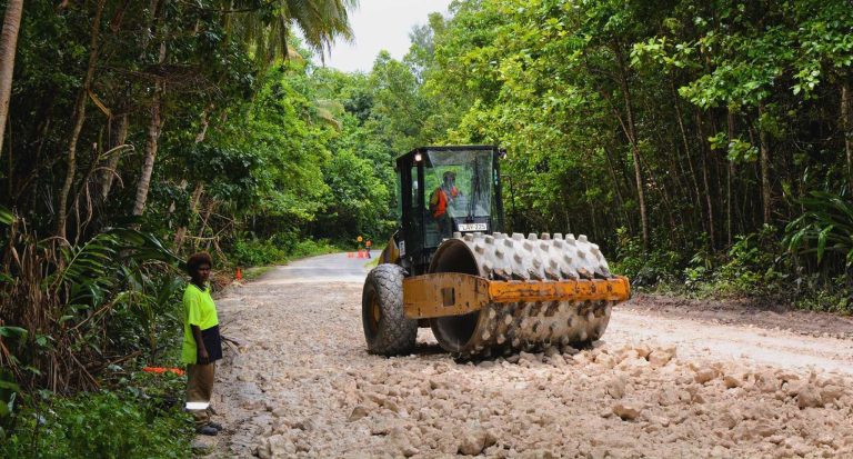 Road construction vehicle compacting a gravel road through dense tropical forest, with workers visible along the roadside.