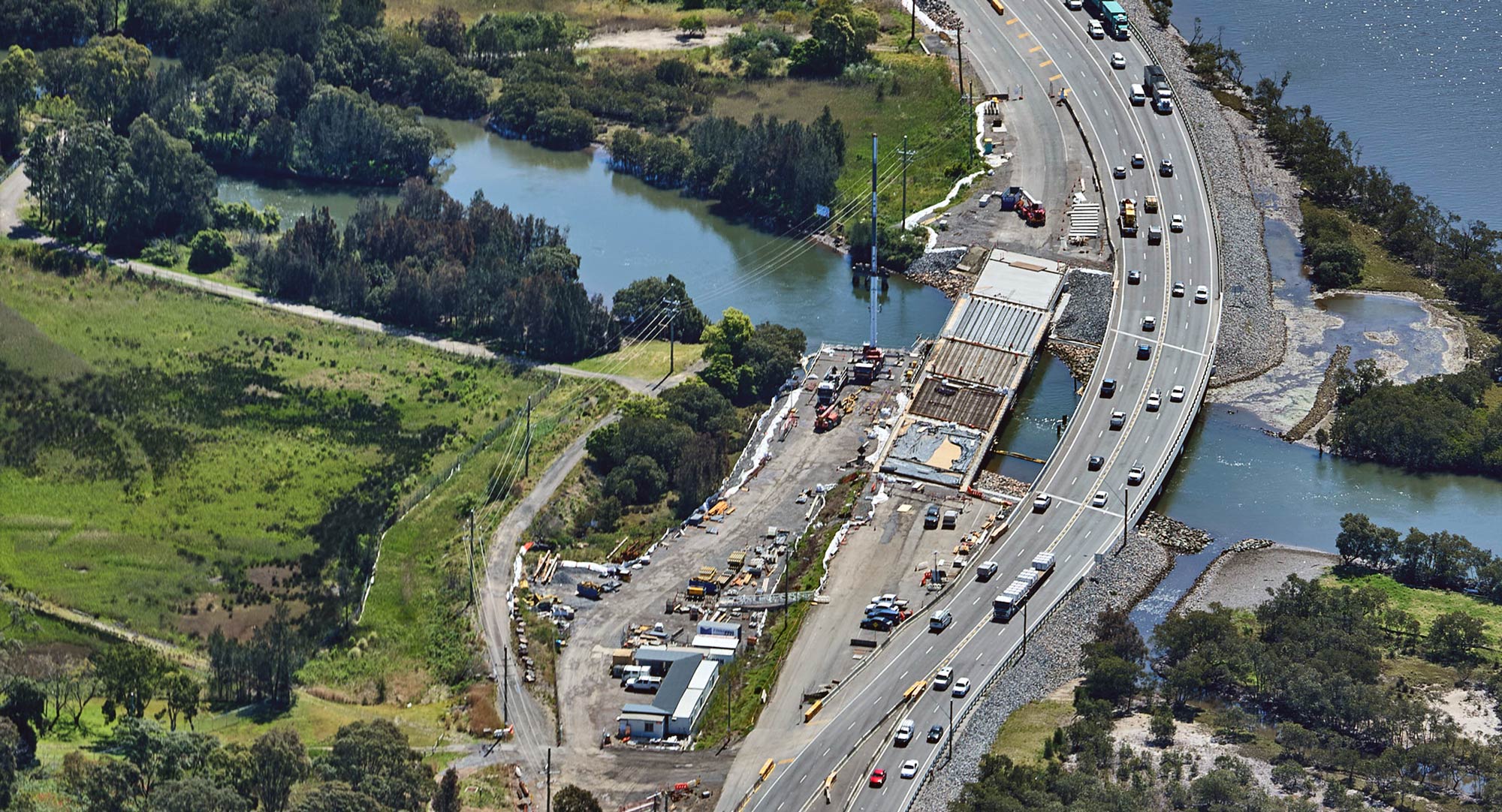 Aerial view of Hexham, showing a widened highway running alongside wetlands and a waterway, with vehicles traveling under overcast skies.