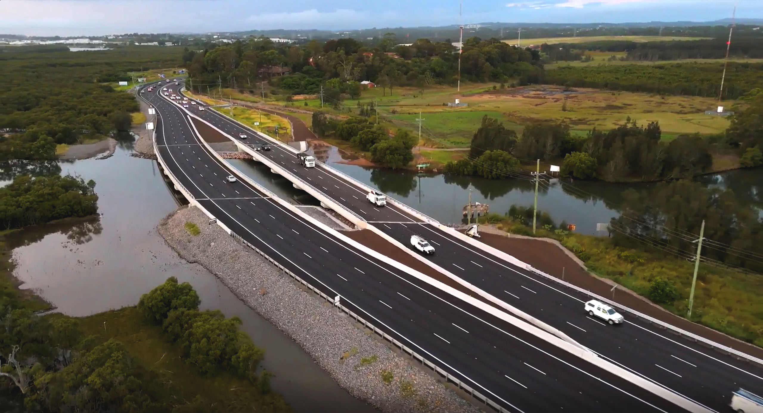 Aerial view of Hexham, showing a widened highway running alongside wetlands and a waterway, with vehicles traveling under overcast skies.