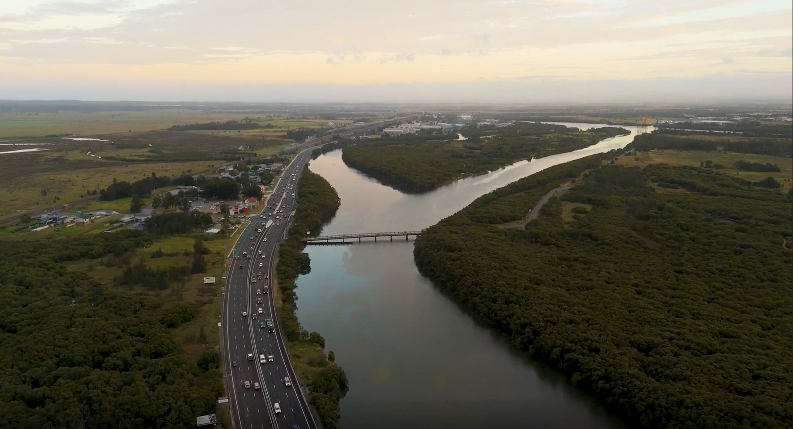 Aerial view of Hexham, showing a widened highway running alongside wetlands and a waterway, with vehicles traveling under overcast skies.