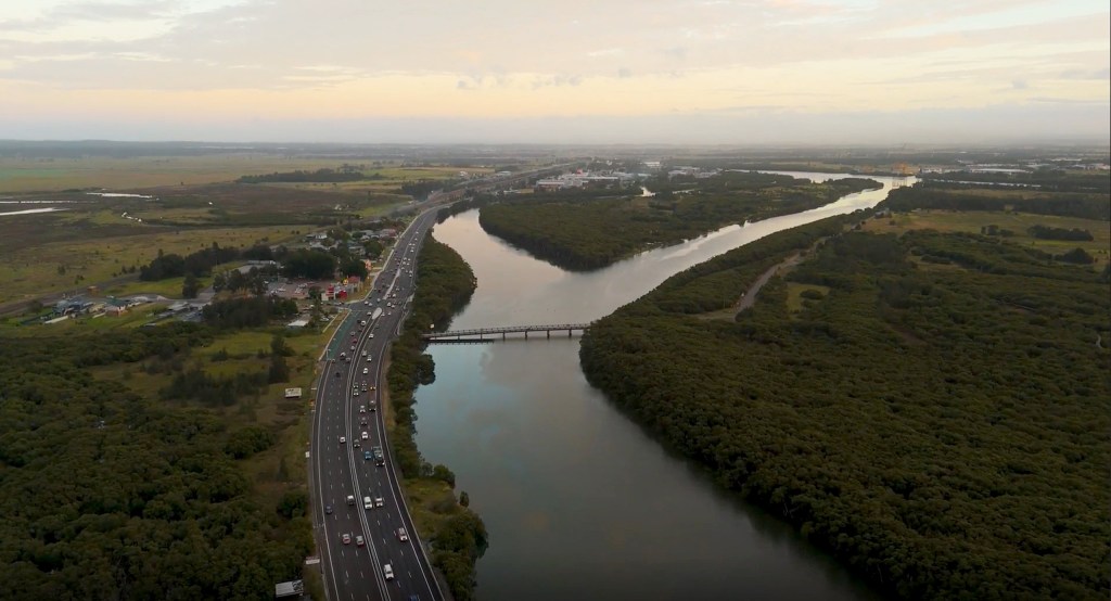 Aerial view of Hexham, showing a widened highway running alongside wetlands and a waterway, with vehicles traveling under overcast skies.