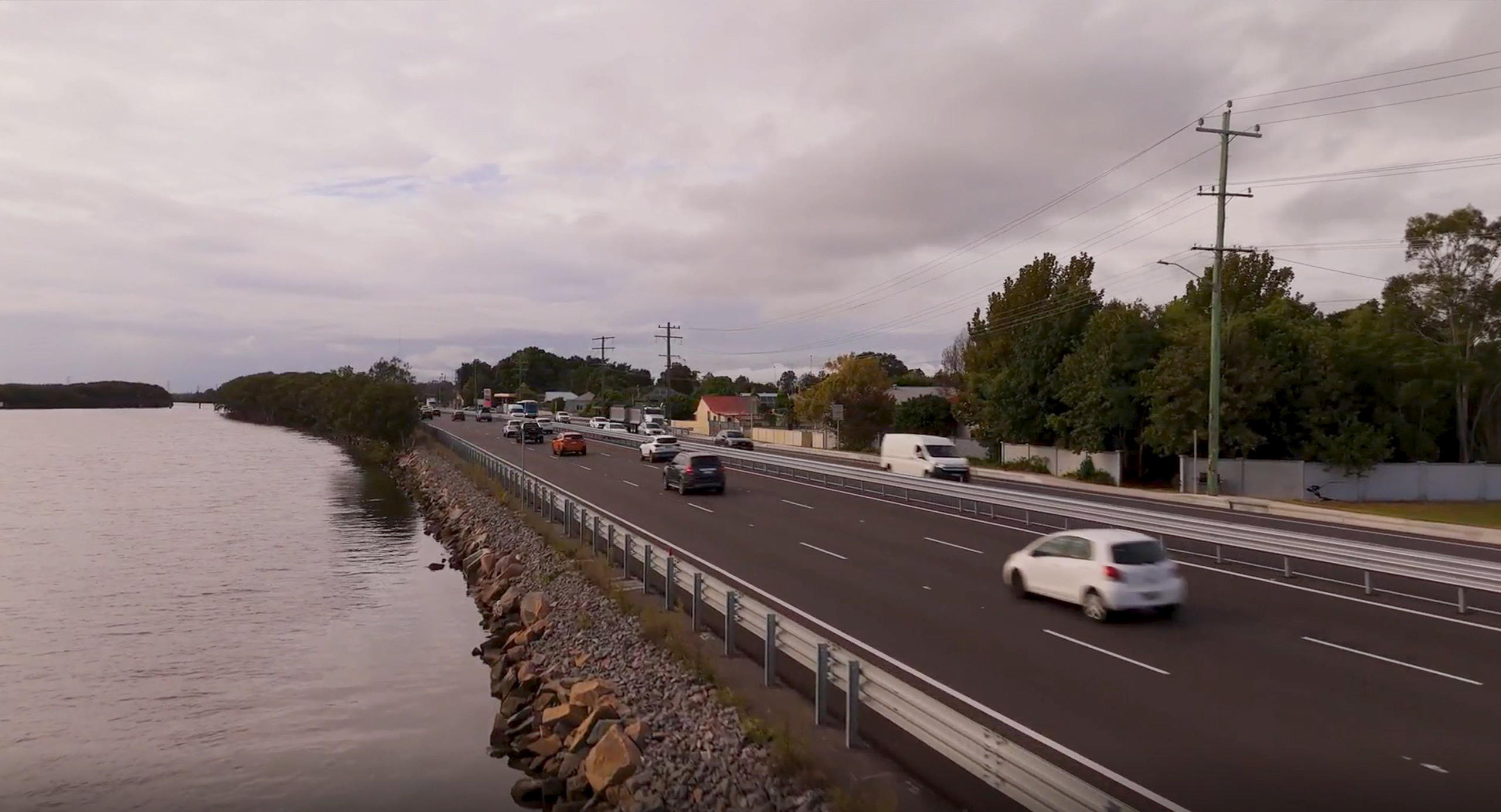 Aerial view of Hexham, showing a widened highway running alongside wetlands and a waterway, with vehicles traveling under overcast skies.