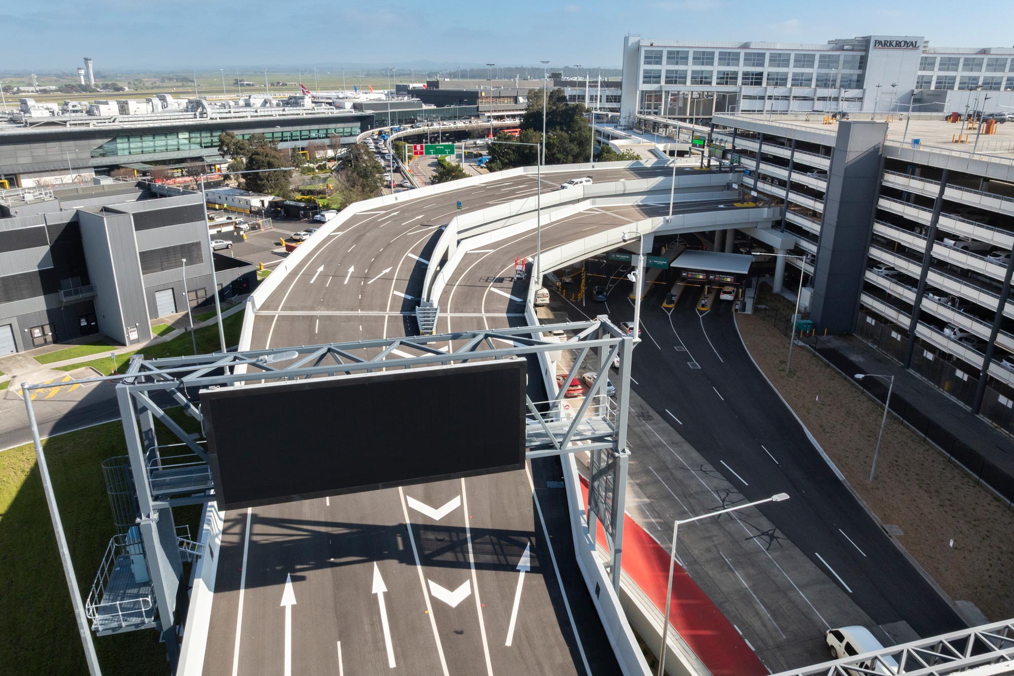 Elevated access roads and airport infrastructure along Melbourne Airport's Naarm Way, with curved multi-lane carriageways, terminal buildings, and a multi-storey car park viewed from above.
