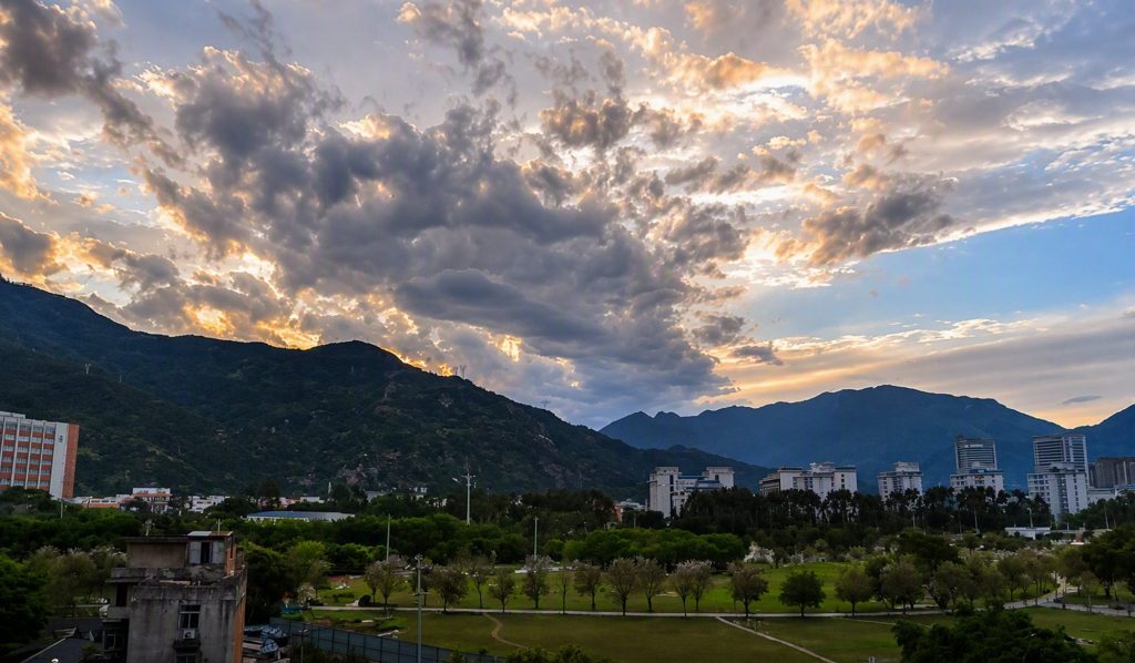 Green urban park and city buildings set against mountains under a dramatic sunset sky with sun rays breaking through dense clouds.