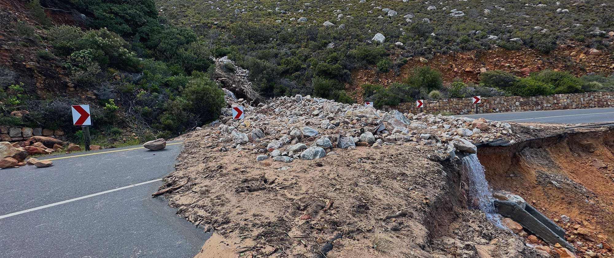Road damaged by landslide and erosion, with rocks and debris blocking part of a mountain roadway.