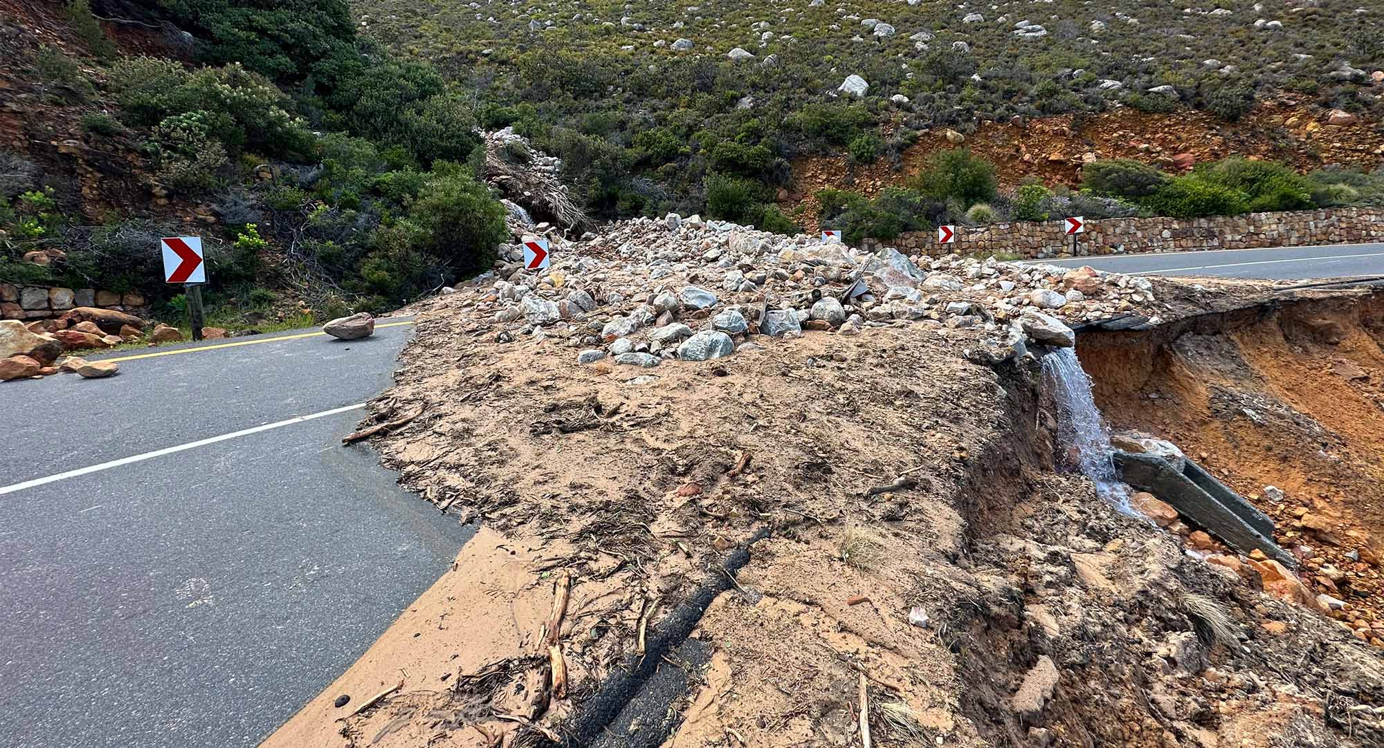Road damaged by landslide and erosion, with rocks and debris blocking part of a mountain roadway.