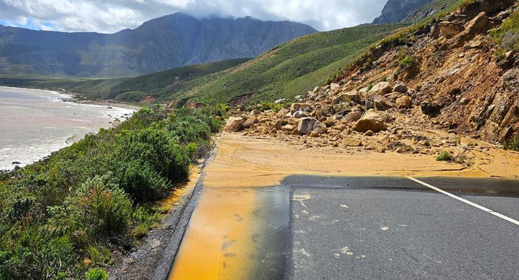Road blocked by a landslide, with rocks and mud covering a coastal roadway beside a steep hillside.