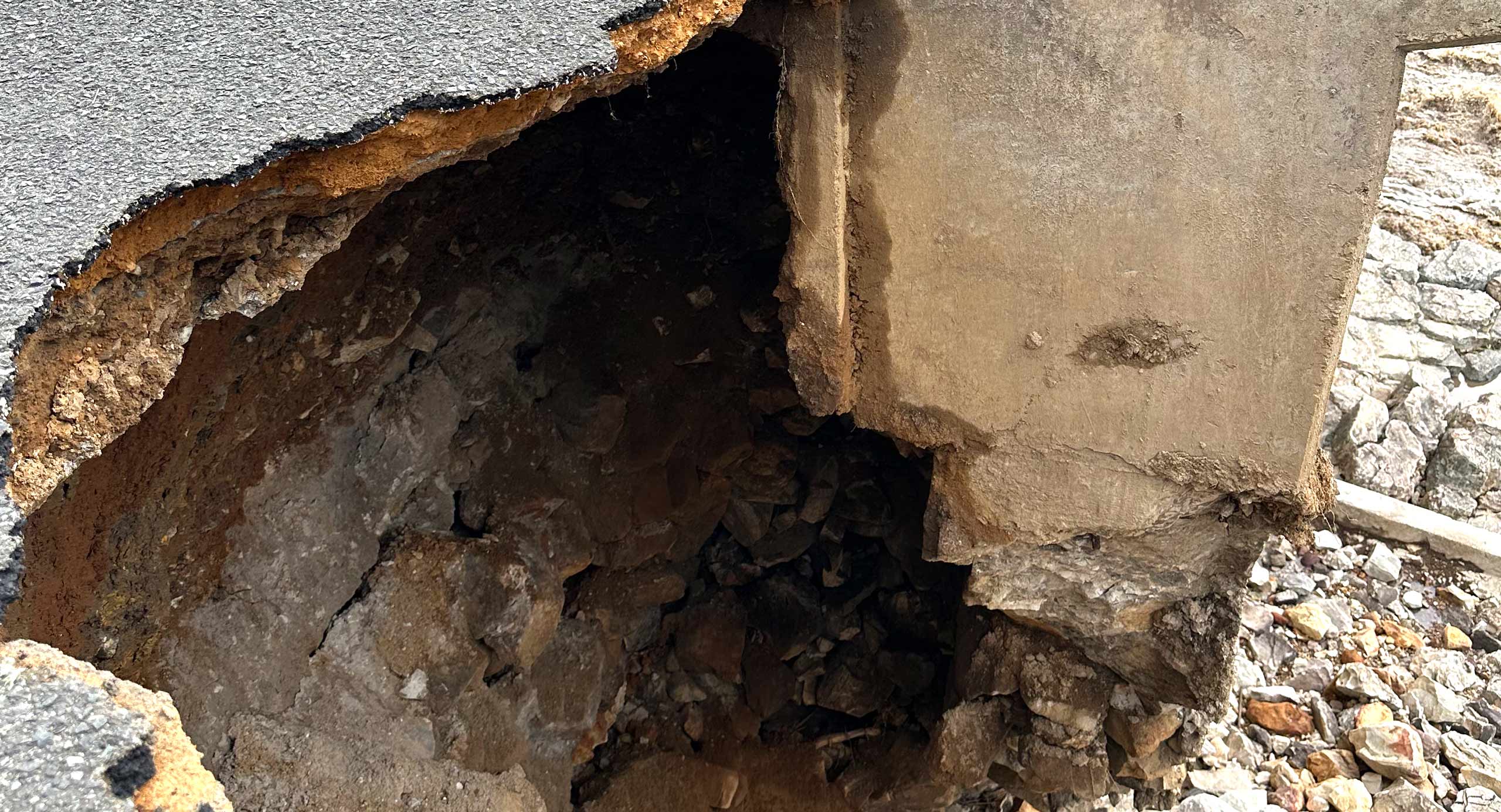 Road damaged by erosion, leaving a large hole beneath the pavement.