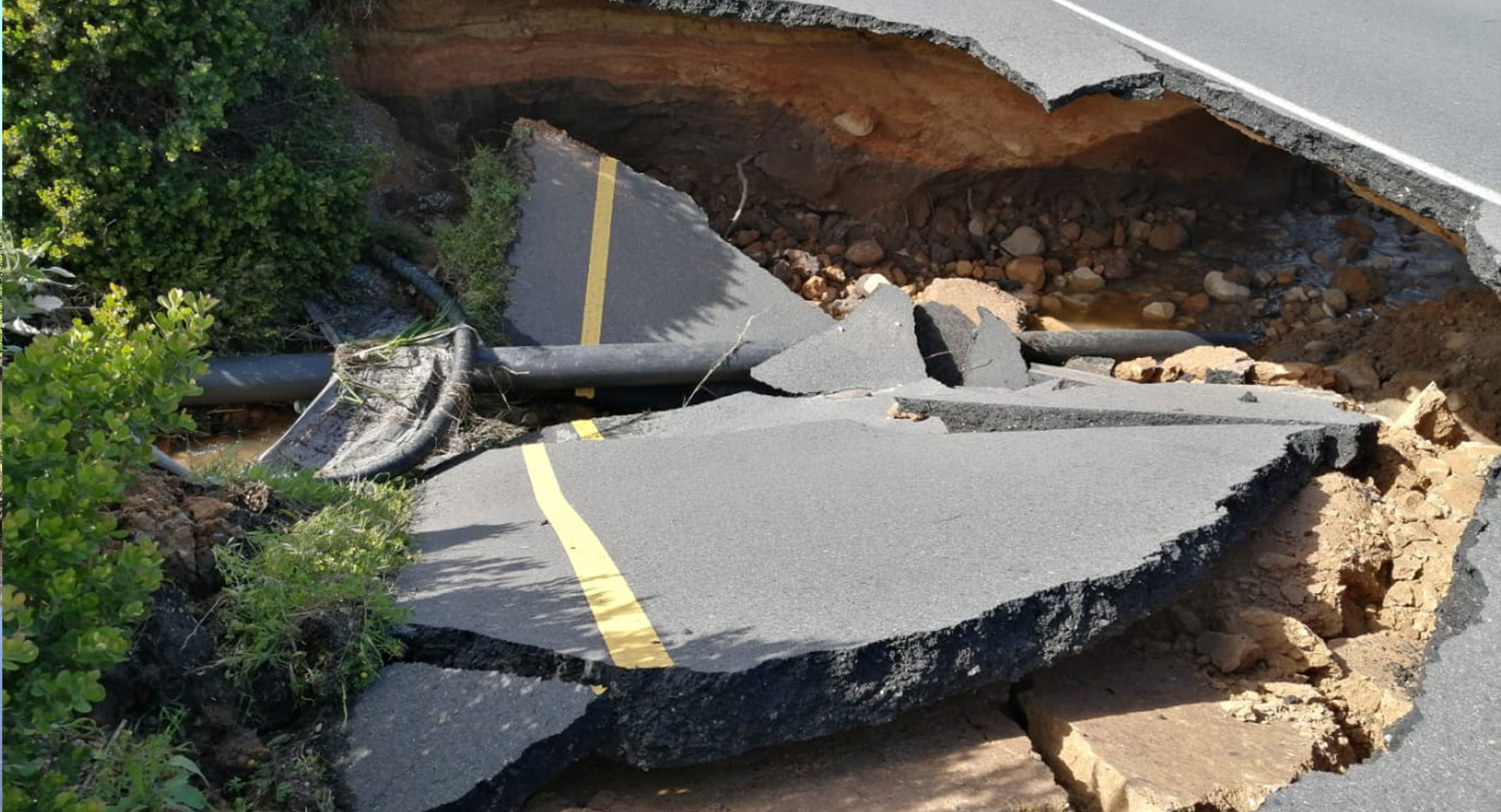 Road collapsed by flood damage, with broken pavement and a large section washed away.